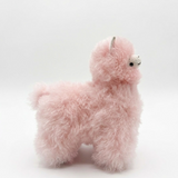 A fluffy light pink alpaca plush toy with small black eyes and a white snout, standing against a plain white studio background.