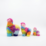 Three fluffy alpaca plush toys in different sizes, each covered in bright rainbow-colored patches of pink, blue, yellow, green, orange, and purple, arranged on a clean white background.