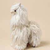 A white alpaca plush toy with long shaggy fur, small black eyes, and tiny black feet, standing against a solid beige background.