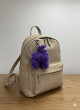 Beige backpack with a purple llama-shaped charm on a wooden surface.