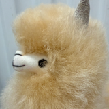 Close-up of a fluffy alpaca plush toy with cream-colored fur, a white snout, and a gray horn, shown against a pale blue-gray background.