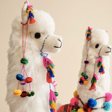 Two large white llama plush toys decorated with colorful tassels, pom-poms, and braided rope headbands, set against a plain beige background.