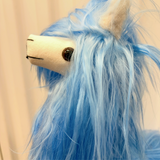 A close-up of a blue fluffy plush toy with a cream-colored snout, embroidered black facial features, and a dark brown bead eye, set against a soft beige background.