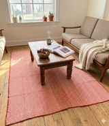 A lifestyle view of Archival Textile No. 110, a vintage Peruvian Frazada rug in dusty rose and peach. The hand-loomed wool rug is styled in a bright, modern living room with a mid-century sofa and wooden coffee table, showcasing its micro-diamond weave as a functional statement area rug.