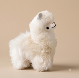 A fluffy white alpaca plush toy with small gray ears and a black stitched face, posed in profile against a warm beige studio background.