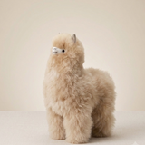 A fluffy  alpaca fur plush toy standing on a light beige surface against a matching neutral background.