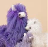 Two fluffy alpaca plush toys posed together against a warm beige background. One large alpaca is purple with a white face, and a smaller white alpaca is nestled beside it.