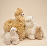 Four fluffy alpaca plush toys in different colors and sizes arranged together on a plain beige studio background.