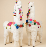 Two live size white alpaca plush toys decorated with colorful tassels, pom-poms, and woven-style saddle blankets, standing against a plain beige studio background.