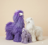 Three fluffy alpaca plush toys in purple and white, arranged together on a light beige surface against a solid warm beige background.