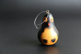 Hand-carved and hand-painted penguin-shaped ornament made from a gourd, with black and orange colors, hanging from a string against a dark background.