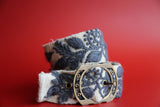 A grey wool belt with hand-embroidered floral patterns and a metallic buckle, displayed against a red background.