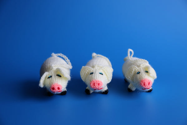 Three hand knitted pig ornaments with white bodies and pink noses, displayed against a blue background.