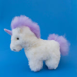 An alpaca plush unicorn toy with fluffy white fur, a purple mane, and a purple tail, standing against a solid blue background.